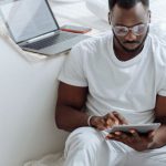 Remote Work - A Man Sitting on the White Blanket on the Floor while Using His Tablet