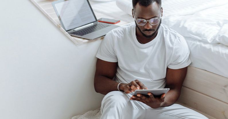 Remote Work - A Man Sitting on the White Blanket on the Floor while Using His Tablet