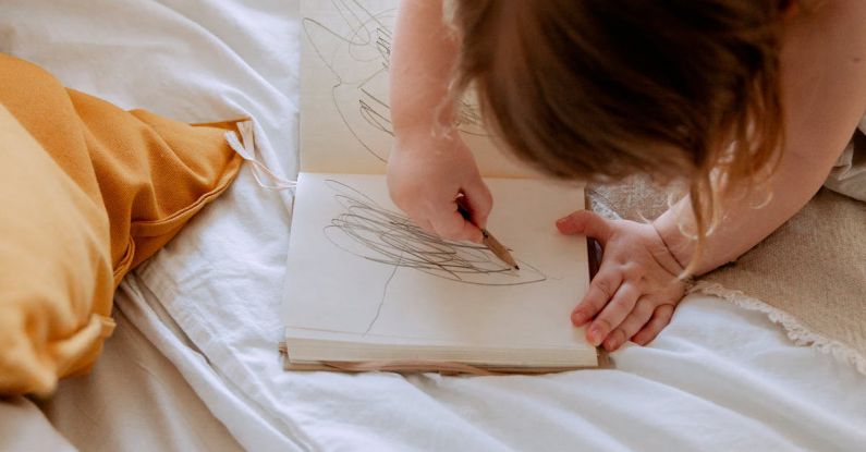 AI Small Business - From above of small girl in dress drawing with pencil in notebook while mother using laptop on bed near