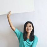 VR Real Estate - Cheerful Asian woman sitting cross legged on floor against white wall in empty apartment and showing white blank banner