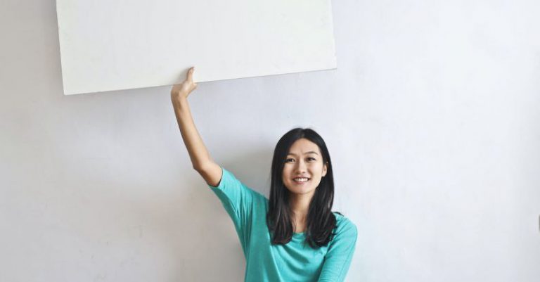 VR Real Estate - Cheerful Asian woman sitting cross legged on floor against white wall in empty apartment and showing white blank banner