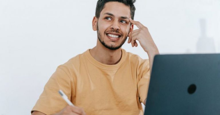 Freelance Future - Smiling young bearded Hispanic male entrepreneur thinking over new ideas for startup project and looking away dreamily while working at table with laptop and taking notes in notebook