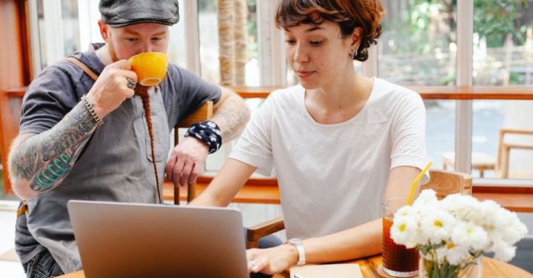 Remote Team Global - Pensive young woman typing on netbook while man with cup drinking coffee at table of cafe