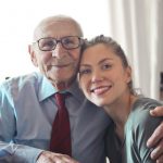 Elderly Care Tech - Positive senior man in formal wear and eyeglasses hugging with young lady while sitting at table