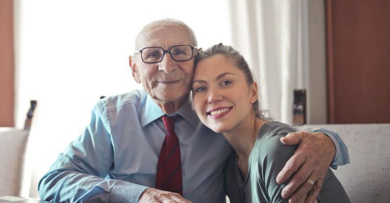 Elderly Care Tech - Positive senior man in formal wear and eyeglasses hugging with young lady while sitting at table