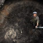 Asteroid Mining - Man in Green Shirt and Black Pants Holding Black and White Stick in Brown Cave